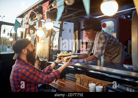 Junge kaukasische bärtige Hipster nimmt zwei Sandwiches von höflich weiblich afroamerikanischer Mitarbeiter im Fast-Food-Service Stockfoto