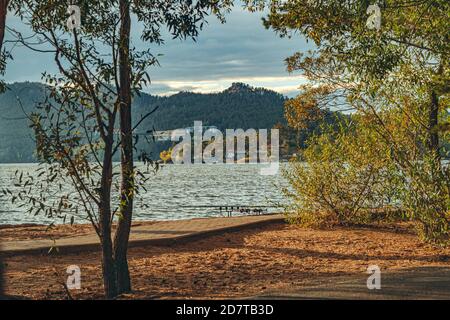 Berg und See durch den Baum im Vordergrund. Landschaft mit See und Bergen gegen den blauen Himmel. Angelruten auf dem See vor dem Hintergrund der Berge. Stockfoto