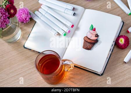 Skizzieren von Cupcake in einem Notizbuch. Alle auf einem Holztisch mit einer transparenten Tasse Tee, Notizbuch, Marker und Blumen platziert.Hobby Skizze. Draufsicht. Weichfokus Stockfoto