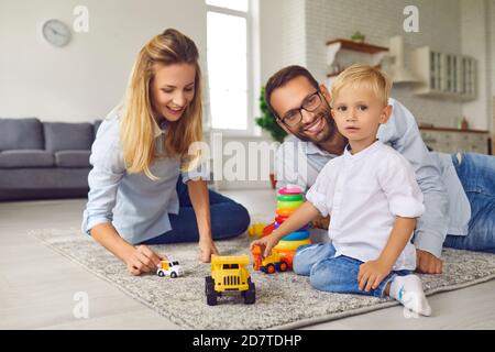 Porträt eines kleinen Jungen, der mit Spielzeugautos spielt, während seine Eltern auf dem Teppich auf dem Boden sitzen. Stockfoto