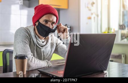 Ein junger Mann sitzt zu Hause vor einem Laptop-Bildschirm, arbeitet aus der Ferne und entfernt eine schützende Gesichtsmaske. Stockfoto