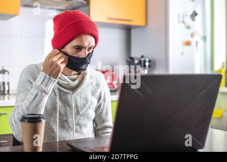 Ein junger Mann sitzt zu Hause vor einem Laptop-Bildschirm, arbeitet aus der Ferne und entfernt eine schützende Gesichtsmaske. Stockfoto