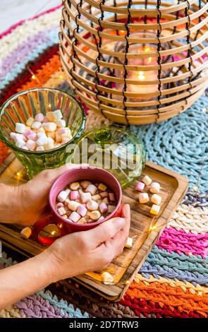 Hände halten rote Tasse Kakao mit pastellfarbenen Marshmallows, Draht und Bambus Laterne mit Kerze auf bunten Häkelteppich aus umfunktionierten T-Shirts. Stockfoto