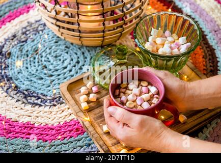 Hände halten rote Tasse Kakao mit pastellfarbenen Marshmallows, Draht und Bambus Laterne mit Kerze auf bunten Häkelteppich aus umfunktionierten T-Shirts. Stockfoto