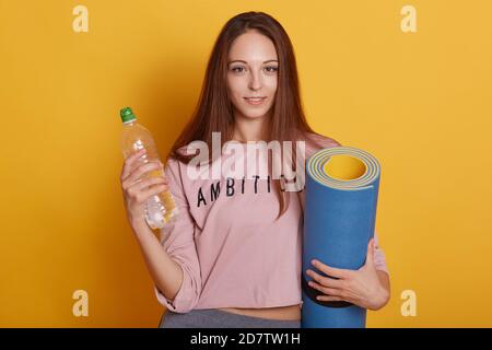 Nahaufnahme Porträt von liebenswert charmante Mädchen mit langen Haaren, posiert isoliert über gelben Studio-Hintergrund, hält Yoga-Matte und eine Flasche Wasser in der Hand Stockfoto