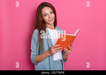 Portrait von positiven süßen jungen Frau mit langen Haaren, trägt blauen Anzug, in guter Stimmung, mit niedlichen Lächeln, halten Notebook in beiden Händen, aussehen Stockfoto