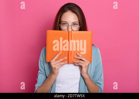 Bild von attraktiven gut aussehenden süßen Buch Wurm bedeckt die Hälfte ihres Gesichts mit Buch, trägt intelligente Kleidung und Accessoires, halten Buch in den Händen, Stockfoto