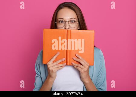 Nahaufnahme Porträt der Student Mädchen versteckt sich hinter Buch, Studio-Foto über rosa Hintergrund, tragen lässige Kleidung und Brille, Buch lesen, halten tex Stockfoto