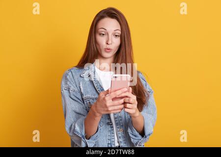 Porträt der attraktiven Caucasasian Frau bekommt überraschende Nachrichten per Telefonanruf, hält die Lippen abgerundet, trägt stilvolle Denim-Jacke, hat Augen aufgetaucht, Stockfoto