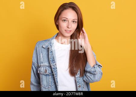 Horizontale Aufnahme von jungen charmanten Frau in Denim Jacke und weißen Casual Shirt stehen über gelben Hintergrund, berührt ihre schöne lange Haare, looki Stockfoto