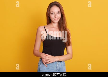 Innenaufnahme einer lebhaften Frau in Jeans und schwarzem T-Shirt Posiert vor der Kamera mit einem angenehmen Lächeln und hält die Hände zusammen Auf Hüfte isoliert über yel Stockfoto