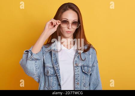 Nahaufnahme Porträt von glamourösen stilvollen Teenager posiert isoliert über gelben Hintergrund im Studio, trägt trendige Sonnenbrille und Denim-Jacke, als al Stockfoto