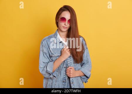 Horizontales Studio-Bild von schönen süßen zarten jungen Frau mit langen Haaren direkt auf die Kamera, tragen trendige Sonnenbrille und Denim-Jacke Stockfoto