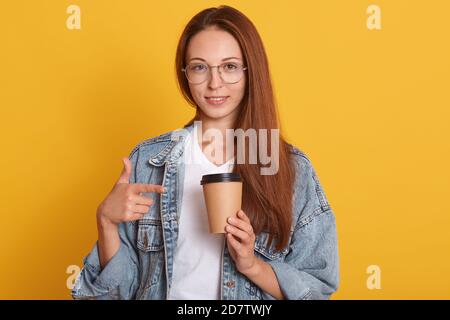 Bild der jungen dunkelhaarigen Frau zeigt auf Papier Kaffee Tasse, die in der Hand hält, Frau gekleidet Denim Jacke und weißen T-Shirt, genießen lecker heiß t Stockfoto