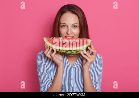 Nahaufnahme Porträt von glücklich schöne junge Frau mit dunklen geraden Haaren essen große Scheibe Wassermelone isoliert über rosa Hintergrund, Dame trägt st Stockfoto