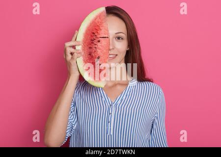 Nahaufnahme Porträt der Frau mit Wassermelone in der Hand, Dame in lässigem Outfit über die Hälfte ihres Gesichts mit Wassermelone, posiert isoliert über pinkstu Stockfoto