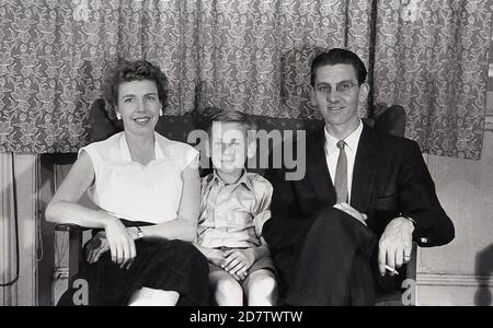 1950s, historisches Familienbild einer Mutter, eines Vaters und eines Sohnes, die auf einem Liegestuhl sitzen. Beide Erwachsenen sind formell angezogen und der Mann trägt einen Anzug mit Krawatte und hat eine Zigarette in der Hand, wie es in dieser Zeit üblich gewesen wäre, da Rauchen für viele Erwachsene die Norm war. Stockfoto