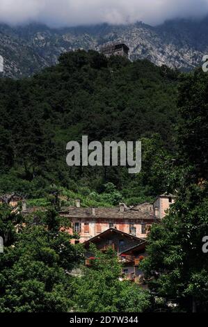 Typische Szene in den Hochebenen nördlich von Pont-Saint-Martin, Donnas und Bard im Aostatal, Italien, mit einem bemalten Bauernhaus mit versperrten Fenstern unter einem traditionellen Steinfischdach durchbohrt von Steinkaminen inmitten von Weinbergen und hinter typischen modernen alpinen Chalets mit blumengeschmückten Holzbalkonen. Hoch über dem Weiler, ein mittelalterlicher Steinturm, unter Restaurierung, Barches auf einer dicht bewaldeten Klippe steigt zu steilen Felsen von treibenden grauen Wolken bedeckt. Stockfoto