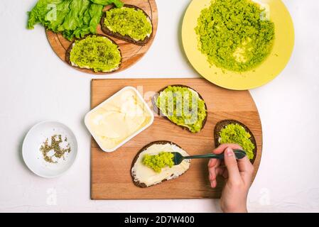 Die Köchin bereitet ein Avocado-Sandwich zu. Tisch mit einem Teller mit Avocado-Püree, Gewürzen, Brot und einer Packung Frischkäse Stockfoto