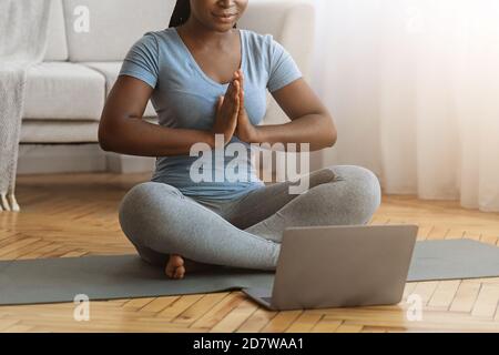 Schwarze Dame meditiert zu Hause, sitzt auf der Matte vor dem Laptop Stockfoto