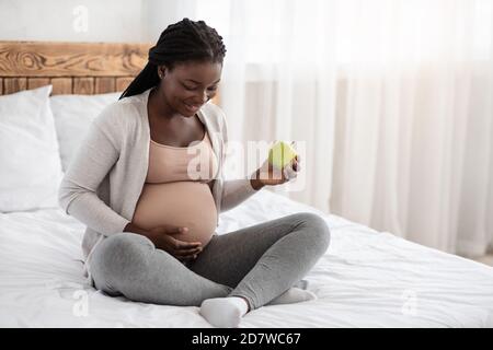 Gesunder Snack. Schwarze schwangere Frau essen grünen Apfel beim Entspannen auf dem Bett Stockfoto