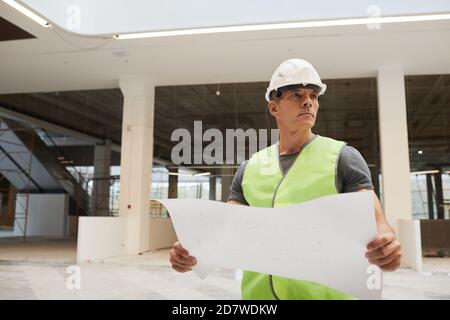 Waist up Porträt von professionellen Bauarbeiter hält Pläne und wegschauen, während im Bürogebäude stehen, kopieren Raum Stockfoto