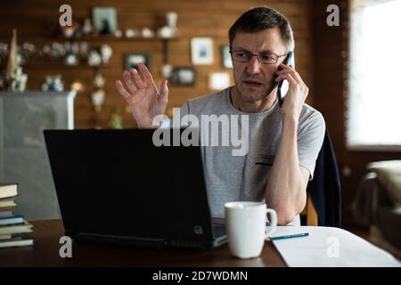 Mann mit mittlerem Erwachsenenalter sitzt vor dem Laptop und spricht am Telefon. Stockfoto