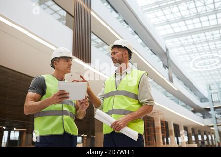 Low-Angle-Porträt von zwei professionellen Bauunternehmen mit digitalen Tablet, während auf der Baustelle im Bürogebäude stehen, Platz kopieren Stockfoto