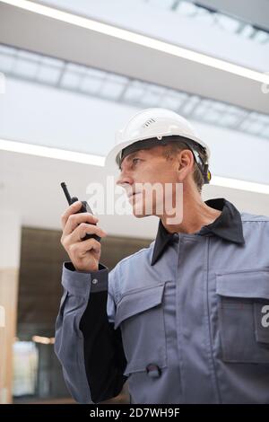Vertikales Low-Angle-Portrait eines reifen Arbeiters, der mit Walkie-Talkie spricht, während er die Arbeit auf der Baustelle oder in der Industriewerkstatt überwacht, Raum kopieren Stockfoto