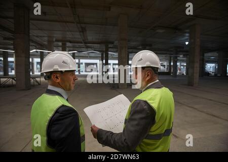 Rückansicht von zwei Geschäftsleuten, die Harthüte tragen und Pläne halten, während sie auf der Baustelle stehen, Platz kopieren Stockfoto