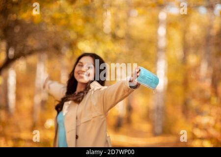 Die Coronavirus-Pandemie ist vorbei. Glückliche kaukasische Frau, die aus chirurgische Maske im Herbst Park, Leerraum. Selektiver Fokus Stockfoto