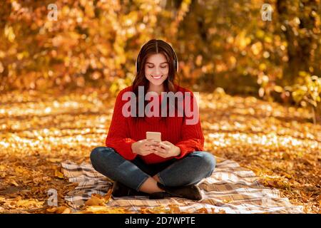 Hübsche junge Frau, die auf einer Picknickdecke im Herbstpark sitzt Mit Kopfhörern und mobilen Geräten und Musik hören Stockfoto