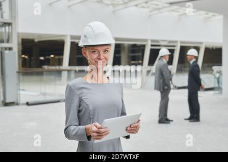 Waist-up-Porträt einer lächelnden Geschäftsfrau mit Hardhut und Tablet, während sie auf der Baustelle im Gebäude steht, Platz kopieren Stockfoto