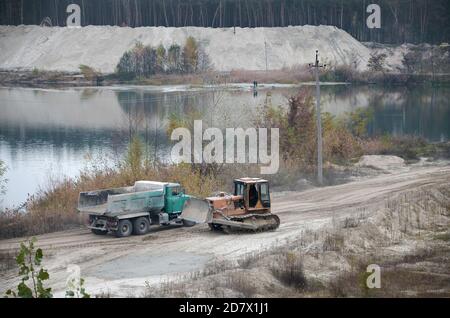 Caterpillar-Lader und Müllkipper arbeiten im Tagebau. Schwere Maschinen im Tagebau am Fluss. Planierraupe und LKW beim Graben und Bagger Stockfoto