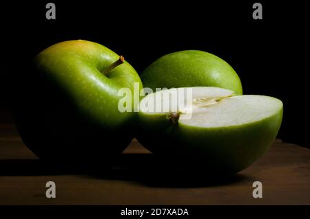 Stillleben, Grüne Apfelhälfte und grüne Äpfel mit einem Chiaroscuro, Barockstil mit schwarzem Hintergrund und auf einem rustikalen Holztisch Stockfoto