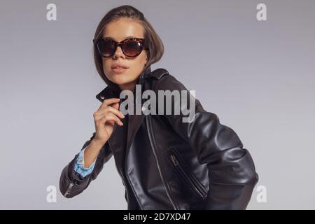 Junge Mode-Modell trägt Lederjacke und Sonnenbrille, auf grauem Studio Hintergrund stehen Stockfoto