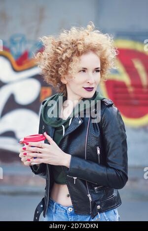 Lockiges rotes Haar Millennial Mädchen tragen in Rock Black Stil im Freien incity in der Nähe von Graffiti Wand. Hipster in Street Fashion Outfit hält Kaffee Tasse. Jugend, Street Style, Lifestyle, Freizeitkonzept. Stockfoto