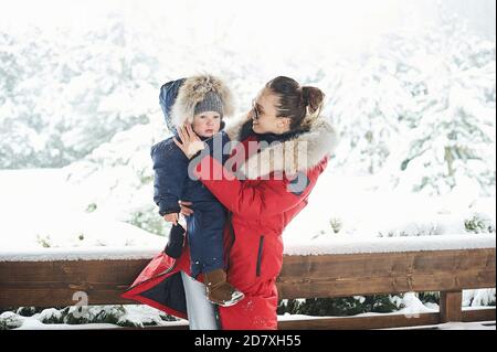 Schöne junge Mutter in roter Jacke mit einem Kind gekleidet Im Winter in der Nähe des Hauses mit einem schneebedeckten weihnachten Baum Stockfoto