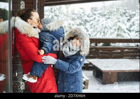 Ein Portret der schönen jungen kaukasischen Mutter mit ihren Kindern Im Winter in der Nähe des Hauses mit einem schneebedeckten weihnachten Bäume im Hintergrund Stockfoto