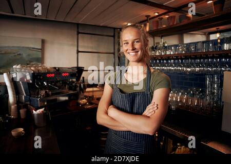 Porträt einer jungen Kellnerin mit Schürze hinter dem Tresen Im Café mit gekreuzten Händen Stockfoto