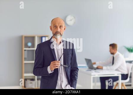 Reifer Unternehmensleiter, der im Büro steht und mit der Kamera schaut Mitarbeiter, der im Hintergrund arbeitet Stockfoto