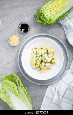 Chinesische Knödel jiaozi in der weißen Platte auf dem grauen Hintergrund mit napa chinakohl und seesamsamen und Soja Sauce mit Serviette Stockfoto