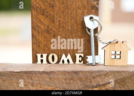 Schlüsselanhänger aus Holz Figur Haus auf Zaun mit Schlüssel und Inschrift in englischen Buchstaben 'Home'. Ferienhaus im Hintergrund. Gebäude, Projekt, Umzug nach n Stockfoto