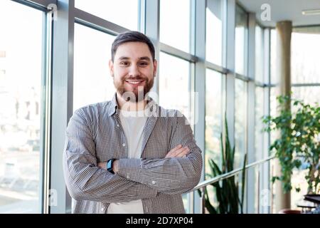 Lächelnder kaukasischer Mann, der allein im Büro an einem großen Fenster stand. Erfolgreicher glücklicher bärtiger junger Mann kreuzte die Arme. Gut aussehender Geschäftsmann Stockfoto