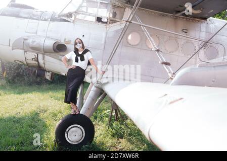 Junge Frau in moderner unifarbener Kleidung, Brille und Maske, die auf einem Vintage-Flugzeug steht und posiert. Stockfoto