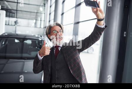 Stilvoller Senior in formeller Kleidung und mit Bart nimmt Selfie gegen moderne Neuwagen. Daumen nach oben zeigen Stockfoto