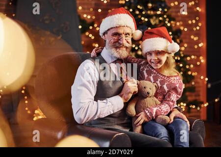 Fröhlicher altmodischer älterer Mann mit grauen Haaren und Bart sitzend Mit kleinen Mädchen in weihnachtsmützen und Teddybär Stockfoto