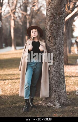 Stilvolle Teenager-Mädchen 14-15 Jahre alt trägt trendigen Wintermantel und Hut posiert im Park lehnen sich auf Baum im Freien. Blick auf die Kamera. Teenagerhood. Stockfoto