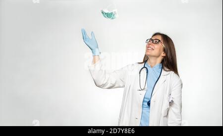 Lächelnde weibliche junge europäische Ärztin wirft Maske weg, nachdem sie erfolgreich das Coronavirus besiegt hat. Stockfoto