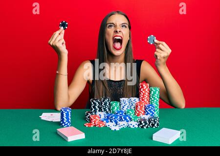 Junge hispanische Frau sitzt auf dem Tisch und hält Casino-Chips wütend und wütend schreiend frustriert und wütend, schreiend mit Wut aufschauend. Stockfoto